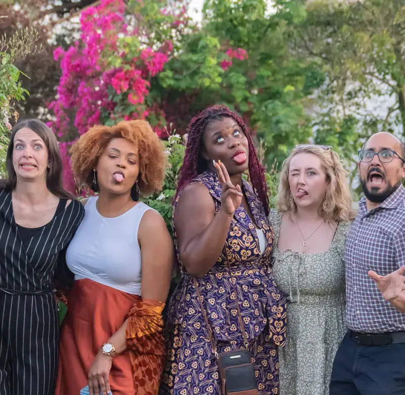 a group of people stand in a line in front of green foliage making silly faces for the camera