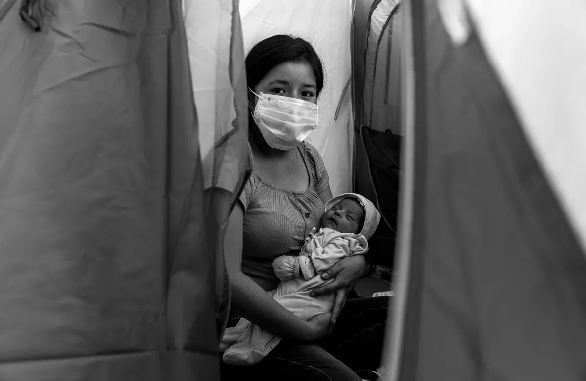 a woman wearing a face mask holds a child in her arms in a makeshift outside structure
